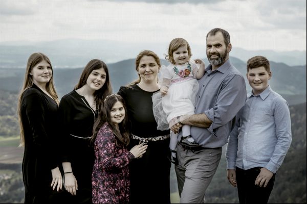 Family photo session Eine glückliche 7-köpfige Familie steht auf dem Berggipfel und hat einen herrlichen Blick auf das grüne Tal unter sich.