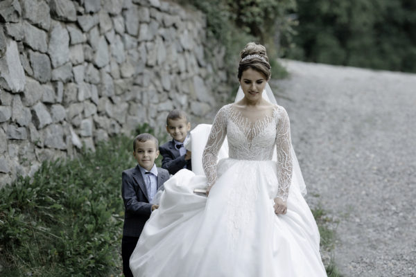 Bride walking by a stone wall, being helped by two little boys holding her wedding dress